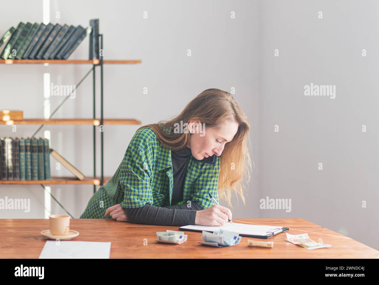 Eine junge hübsche Dame spricht am Telefon und schreibt auf einem Tablet vor dem Hintergrund einer Arbeitsumgebung und Geld auf dem Tisch. Stockfoto