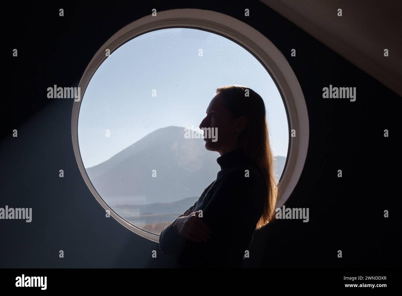 Porträt einer Frau im Profil vor dem Hintergrund eines runden Fensters, hinter dem man die Berge sehen kann. Stockfoto