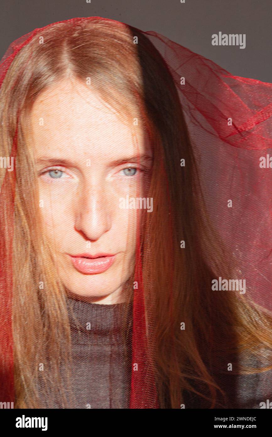 Vertikale Zusammensetzung. Porträt einer erwachsenen jungen Frau mit langen blonden Haaren und grauen Augen in den hellen Sonnenstrahlen und einem roten durchsichtigen Tuch auf Stockfoto
