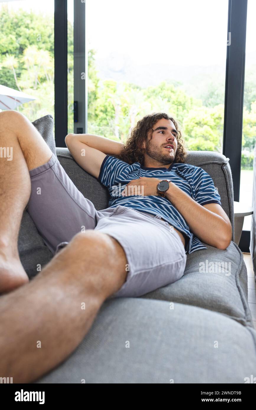 Junger kaukasischer Mann mit lockigen Lounges auf einem Sofa, nachdenklich mit Kopierraum Stockfoto