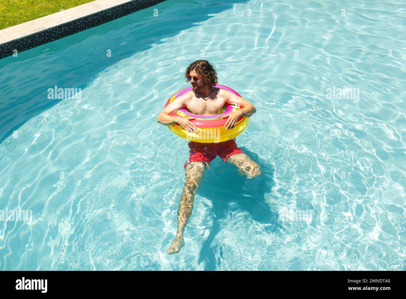 Junger Kaukasier schwimmt in einem Pool mit einem bunten Ring, trägt Sonnenbrille und rote Shorts Stockfoto