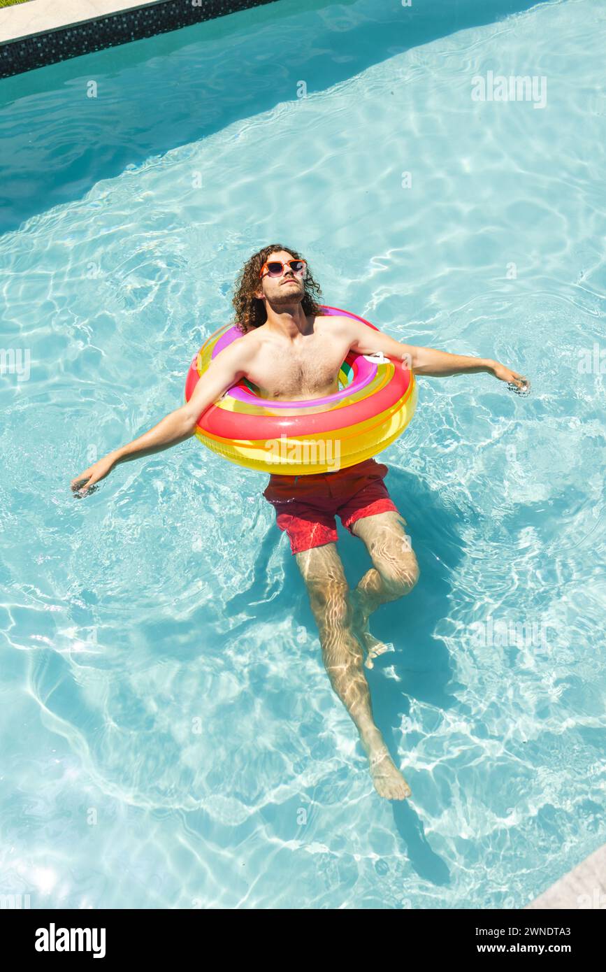 Junger kaukasischer Mann mit lockigen Haaren entspannt sich auf einem farbenfrohen Schwimmer in einem Pool mit Kopierraum Stockfoto