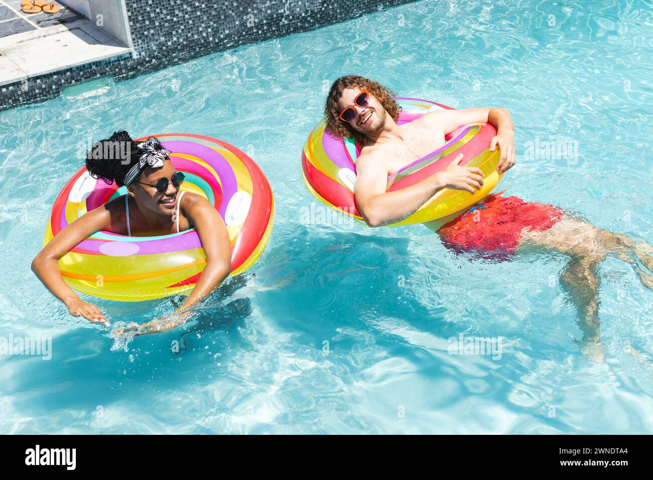Die junge afroamerikanische Frau und der kaukasische Mann genießen einen sonnigen Pooltag, der auf bunten Ringen schwimmt Stockfoto
