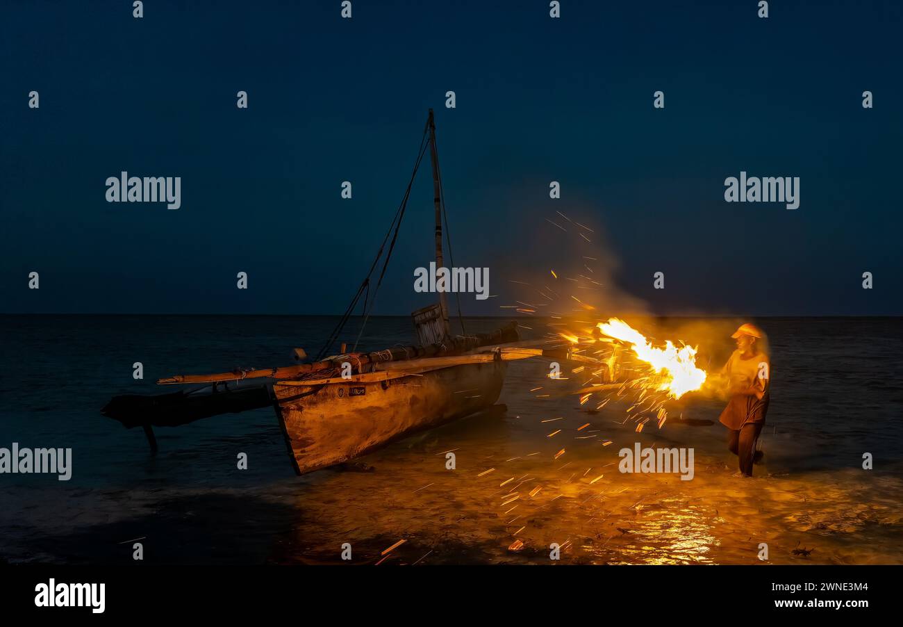 Ein Mann benutzt brennende Palmwedel in der Nacht, um sein traditionelles hölzernes Dhow-Boot, Jambiani, Sansibar, Tansania, zu sairieren und zu wasserdicht zu machen Stockfoto