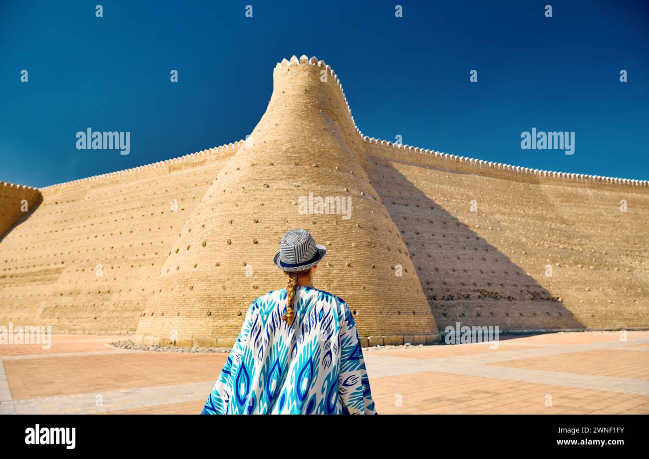 Frau in ethnischer Kleidung mit blauem traditionellem Muster in der Nähe der Mauern der Arche Buchara in Usbekistan. Stockfoto
