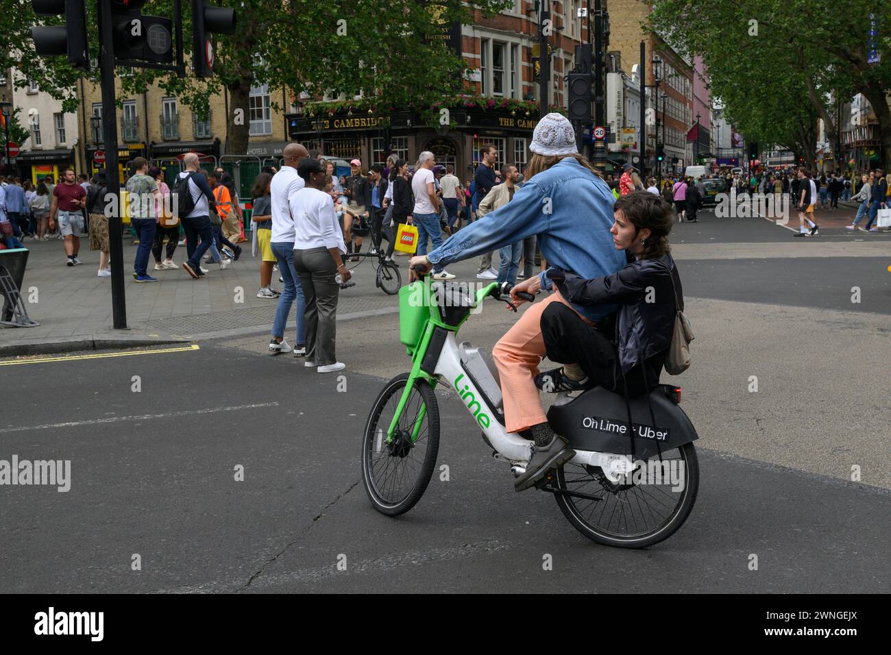 Ein Mann und eine Sozius-Passagierin, die mit einem Lime-Elektro-Mietrad über Cambridge Circus, Charing Cross Road, London, Großbritannien, fahren. Juli 2023 Stockfoto