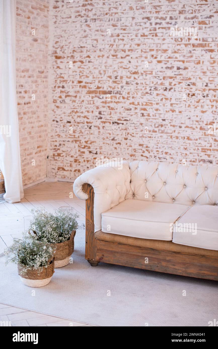 Inneneinrichtung mit einem weißen gesteppten Sofa mit Holzsockel vor einer leeren Ziegelwand, mit weißen Vorhängen und Blumentöpfen auf einem grauen Teppich Stockfoto
