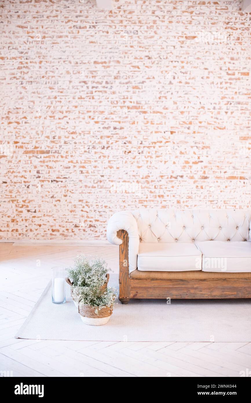 Inneneinrichtung mit einem weißen gesteppten Sofa mit Holzsockel vor einer leeren Ziegelwand, mit weißen Vorhängen und Blumentöpfen auf einem grauen Teppich Stockfoto