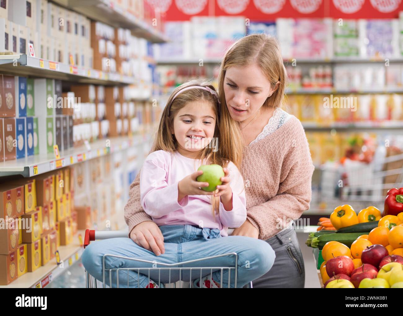 Glückliches süßes Mädchen, das auf einem Einkaufswagen sitzt und frisches Gemüse und Obst mit ihrer Mutter im Supermarkt kauft Stockfoto