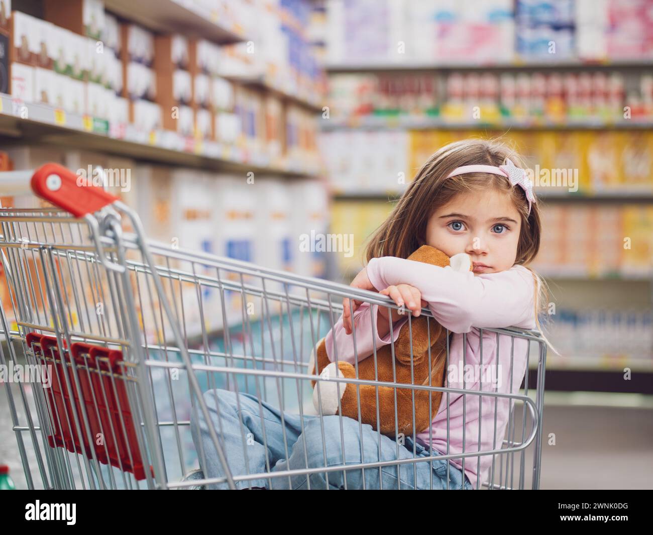 Porträt eines süßen kleinen Mädchens im Supermarkt, sie sitzt in einem Einkaufswagen und umarmt ihren Teddybären Stockfoto