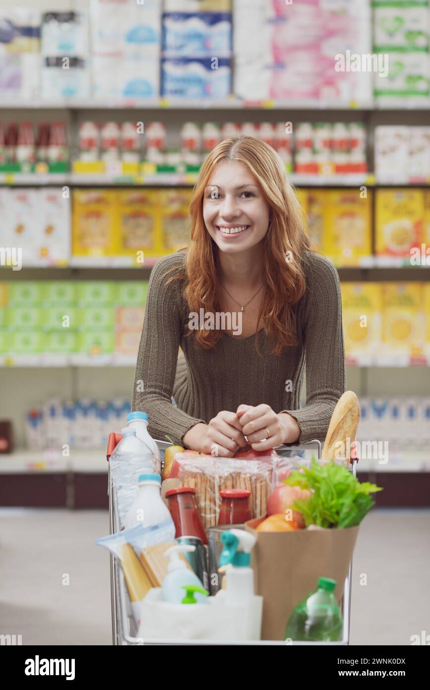 Porträt einer jungen Frau im Supermarkt, sie lehnt sich auf den Griff des Einkaufswagens und lächelt in die Kamera Stockfoto