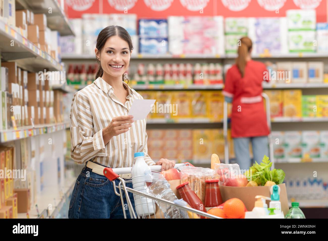 Porträt einer jungen lächelnden Frau, die Lebensmittel im Supermarkt kauft, hält eine Einkaufsliste und schiebt einen vollen Warenkorb Stockfoto