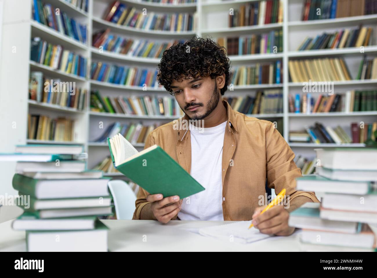 Ein engagierter Student, der in das Lesen eines Buches vertieft ist, umgeben von Haufen von Lehrbüchern, und sich fleißig auf anstehende Prüfungen in der ruhigen Atmosphäre einer Universitätsbibliothek vorbereitet. Stockfoto