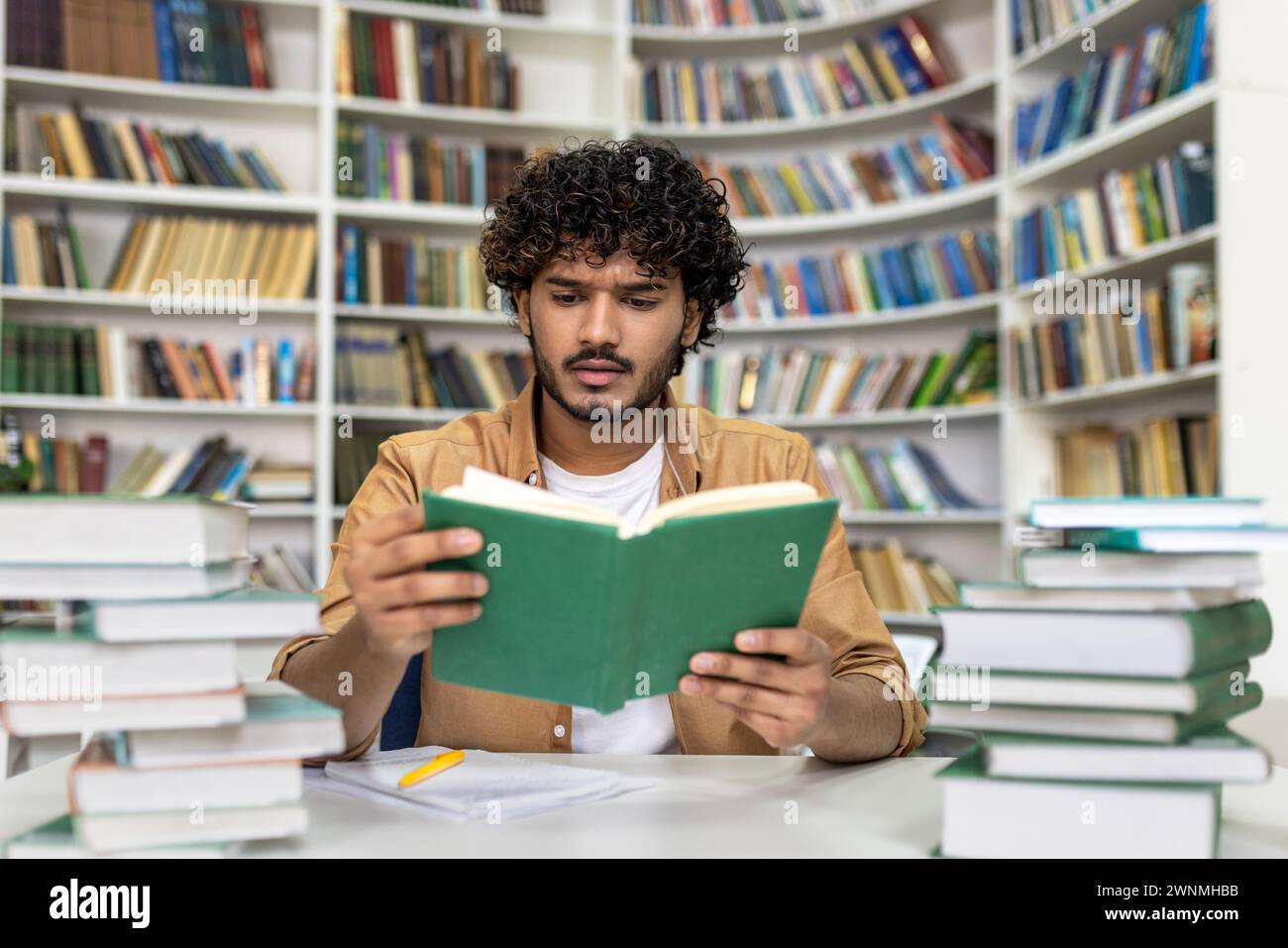 Ein fleißiger junger Mann, der tief in das Lesen eines Buches vertieft ist, ist umgeben von riesigen Literaturstapeln in einer hell beleuchteten Universitätsbibliothek. Das konzentrierte Ambiente unterstreicht die Ernsthaftigkeit der Prüfungsvorbereitung und des akademischen Engagements. Stockfoto
