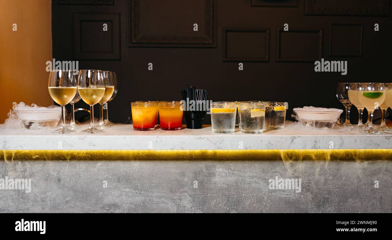 Eine Auswahl an verschiedenen Getränken, darunter Wein, bunte Cocktails und Wasser an einer Bar mit Trockeneis, die einen nebeligen Effekt erzeugt. Stockfoto