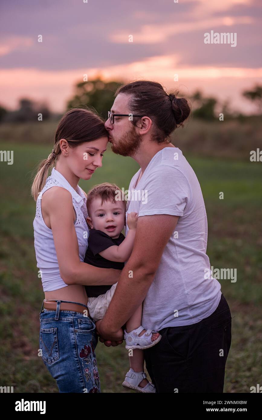 Vielfalt stilvoller Vater küsst Mütter in die Stirn, während er den kleinen Sohn hält, Konzept der schönen Familienverbindung vor dem pulsierenden Sonnenuntergang. Familienbande, Stockfoto