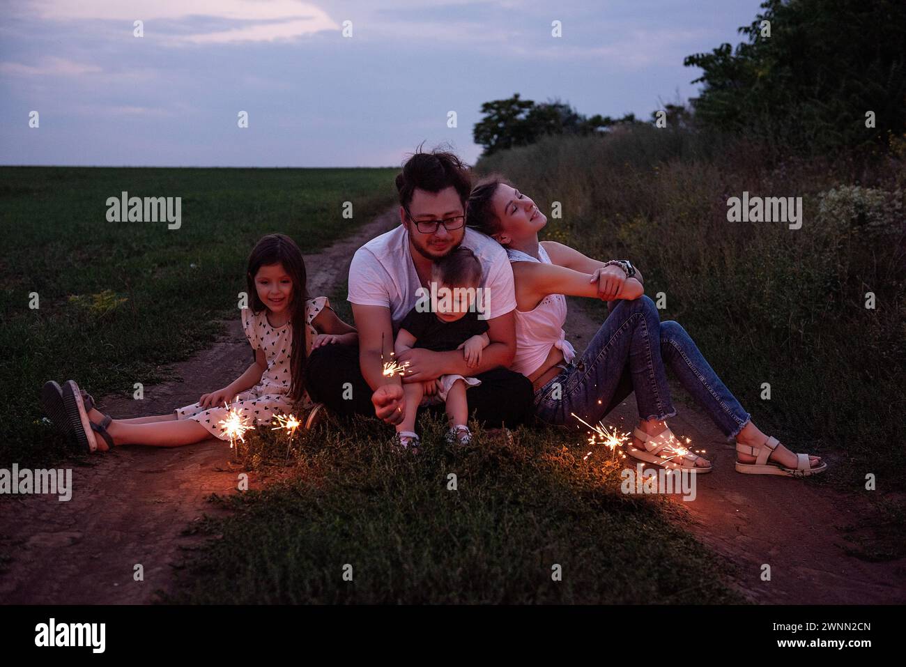 Die vierköpfige Familie spielt mit Funkeln auf dem ländlichen Weg und teilt gemütliche Momente, während die Dämmerung sich einsetzt. Helles Feiern in der Natur Stockfoto