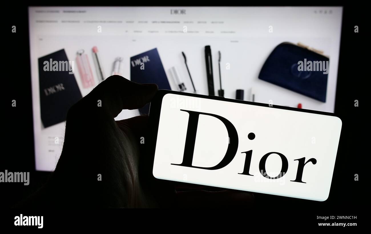 Person, die ein Mobiltelefon mit dem Logo des französischen Luxusmodeunternehmens Christian Dior SE vor der Business-Website hält. Schwerpunkt auf dem Display des Telefons. Stockfoto