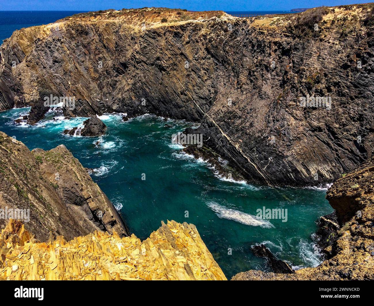 Das Bild zeigt eine zerklüftete Küstenlandschaft mit steilen Klippen, die eine kleine Bucht mit türkisfarbenem Meer umgeben. Das Meer ist turbulent mit weißen Wellen, die sich in Crashin tummeln Stockfoto