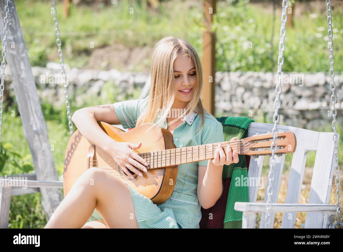 Eine junge Frau sitzt auf der Schaukel und spielt Gitarre im Sommerpark. Blondes weibliches Model mit Gitarre. Mädchen spielt Gitarre im Freien. Stockfoto