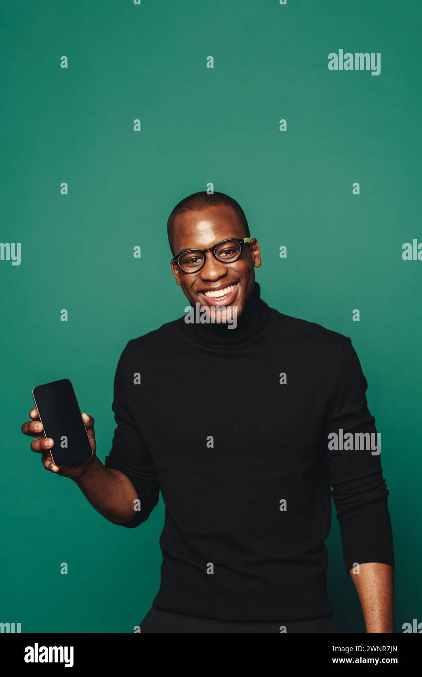 Ein junger Mann, der stilvoll in legere Kleidung gekleidet ist, hält ein trendiges Mobiltelefon vor einem leuchtend grünen Hintergrund. Mit selbstbewusstem und fröhlichem Verhalten, er Stockfoto