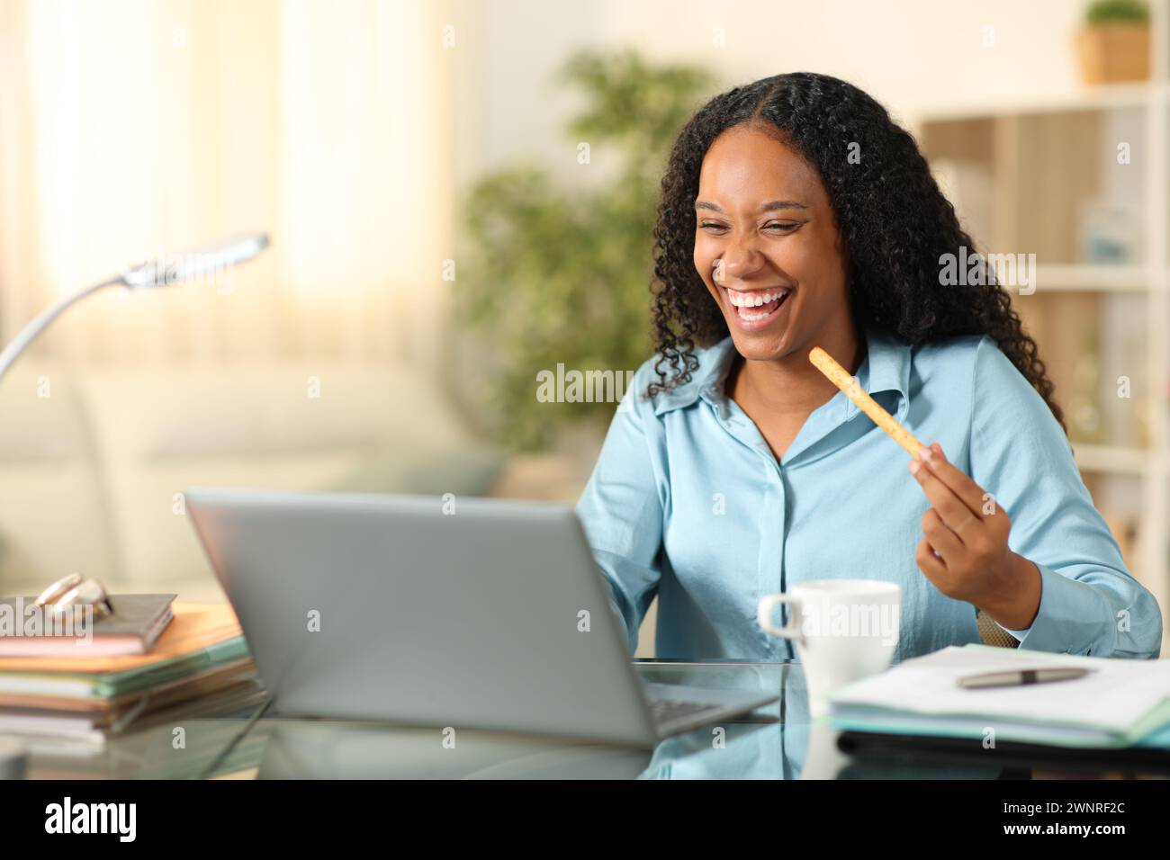 Glückliche schwarze Geschäftsfrau, die zu Hause mit dem Laptop einen Snack isst Stockfoto