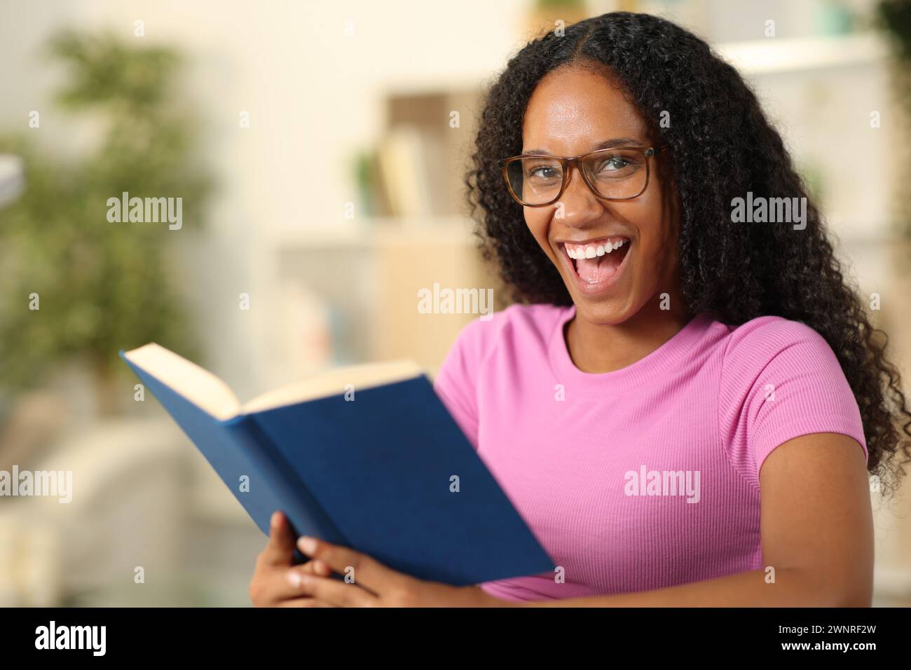 Ein glücklicher schwarzer Leser mit Brille sieht dich an, der ein Papierbuch zu Hause hält Stockfoto