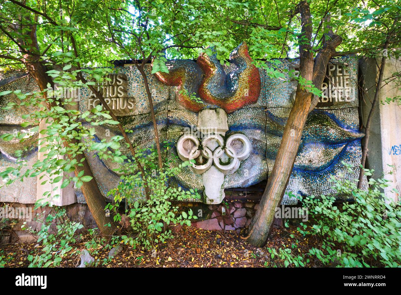 Hinter dem Überwuchs versteckt sich eine Mosaikfliese mit olympischer Flamme, ein Fackelsymbol, das in einen Zaun eingebaut ist. In Almaty, Kasachstan. Stockfoto