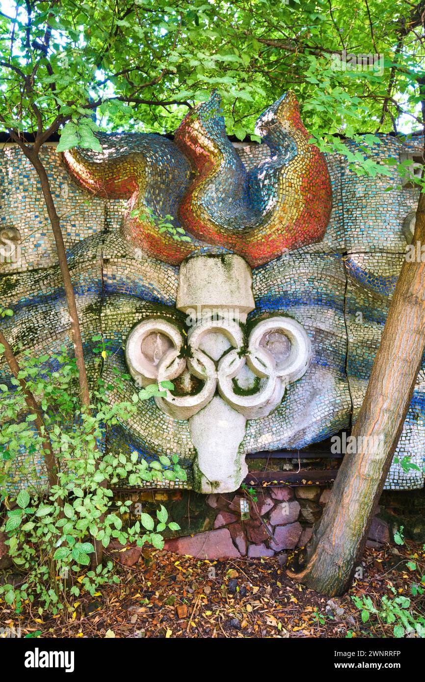 Hinter dem Überwuchs versteckt sich eine Mosaikfliese mit olympischer Flamme, ein Fackelsymbol, das in einen Zaun eingebaut ist. In Almaty, Kasachstan. Stockfoto