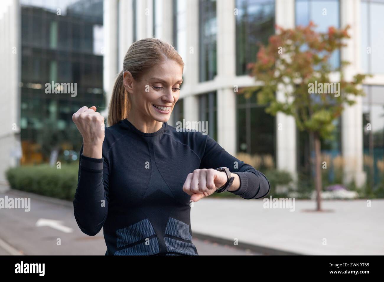 Eine fitte Frau mit einem Lächeln überprüft ihren Fitness-Tracker im Freien und strahlt Glück und einen gesunden Lebensstil aus. Vor dem Hintergrund der urbanen Architektur spiegelt dieses Bild das Wesen des aktiven Lebens in der Stadt wider. Stockfoto