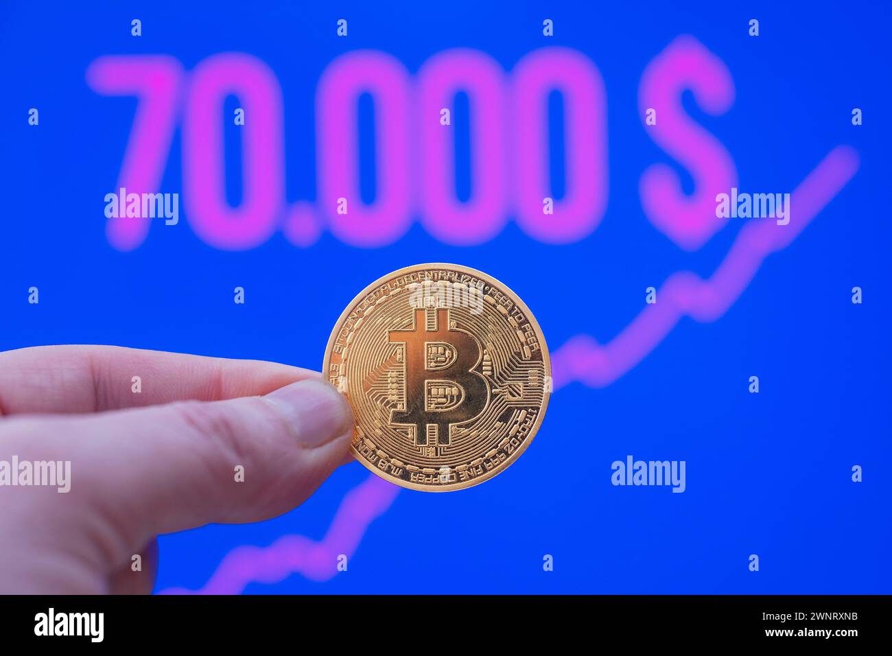 Bitcoin-Münze zwischen den Fingern vor einem Marktpreis von über 70.000 USD Stockfoto