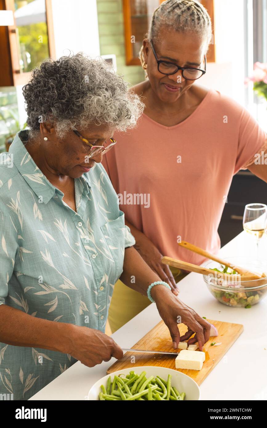Eine ältere afroamerikanische Frau hackt Gemüse, während eine hochrangige birassische Frau mit einem Lächeln zusieht Stockfoto