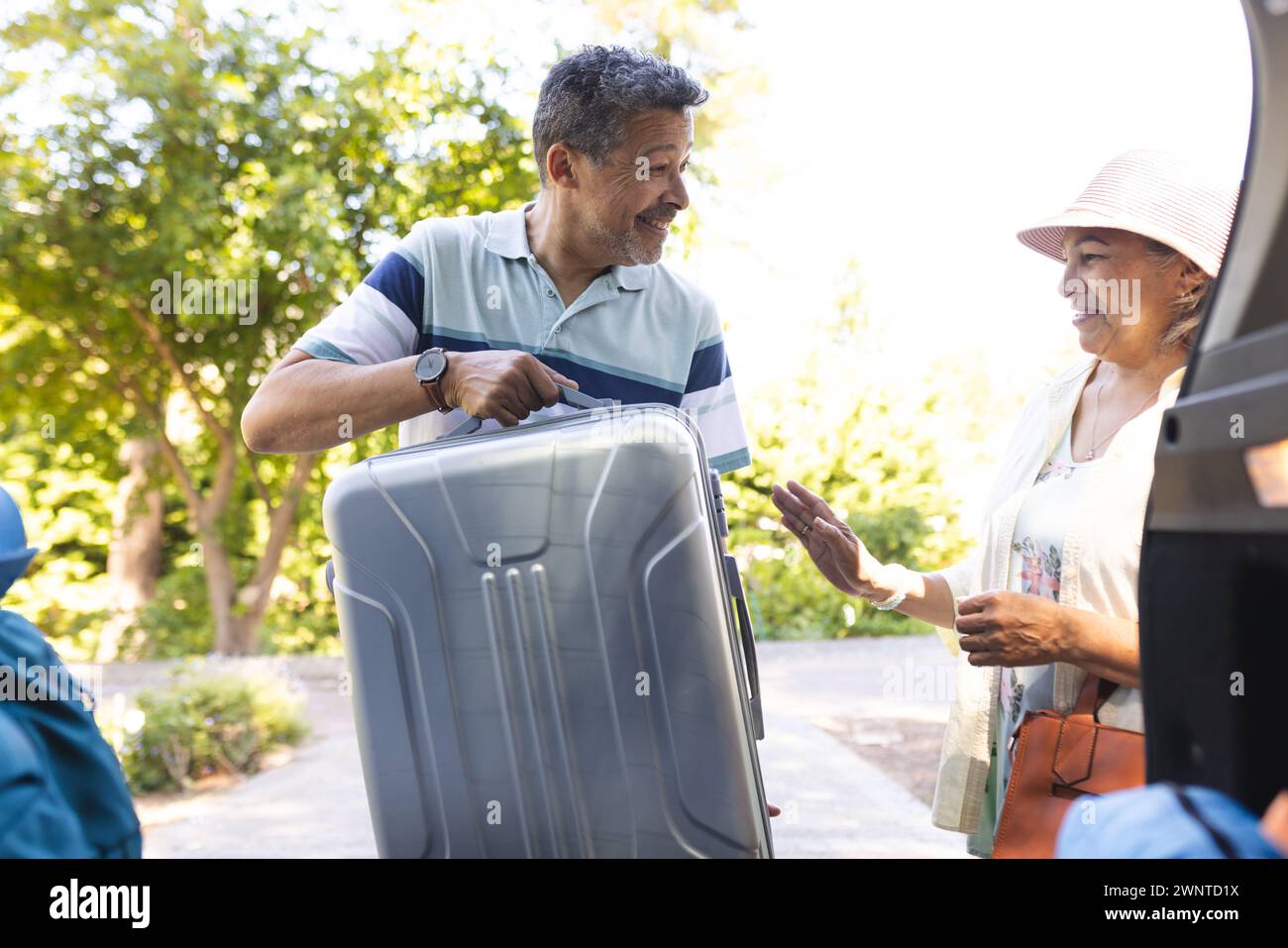 Biracial man hilft älteren Biracial Frau mit Gepäck, beide lächeln in einer sonnigen Umgebung im Freien Stockfoto