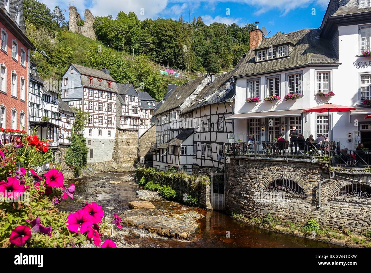 Malerisches europäisches Dorf (Monschau, Deutschland) mit Fachwerkhäusern und einer blumengesäumten Brücke über einen Bach mit grünen Hügeln im Hintergrund Stockfoto