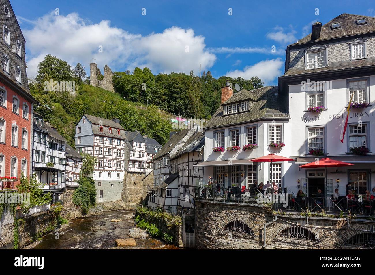 Malerisches europäisches Dorf (Monschau, Deutschland) mit Fachwerkhäusern und einer blumengesäumten Brücke über einen Bach mit grünen Hügeln im Hintergrund Stockfoto