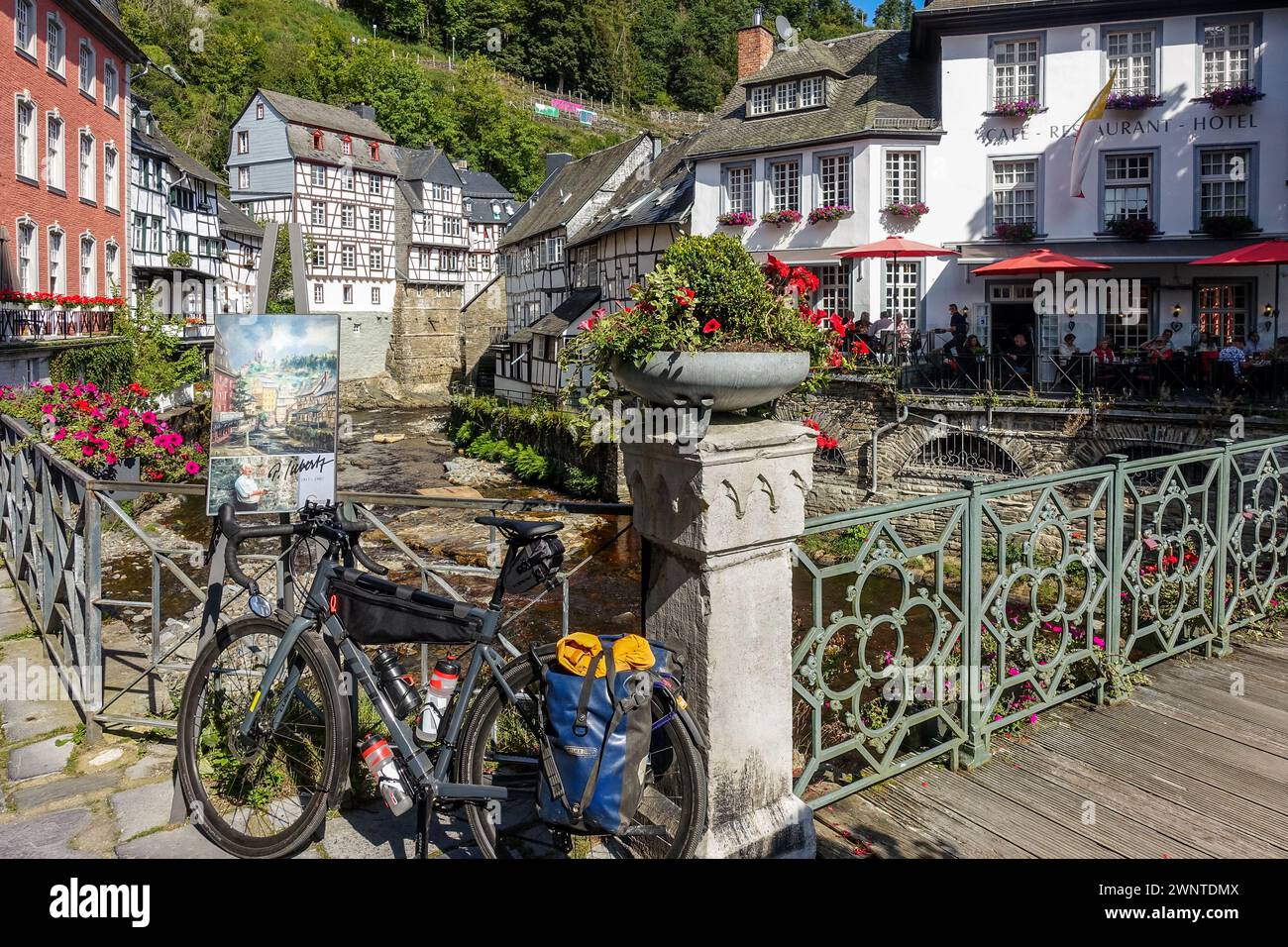 Schotterrad in einem malerischen europäischen Dorf (Monschau, Deutschland) mit Fachwerkhäusern und einer blumengesäumten Brücke über einen Bach Stockfoto