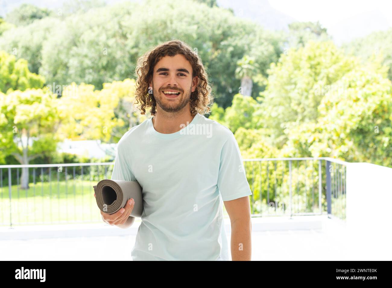 Junger Kaukasier mit lockigen Haaren hält draußen eine Yogamatte Stockfoto