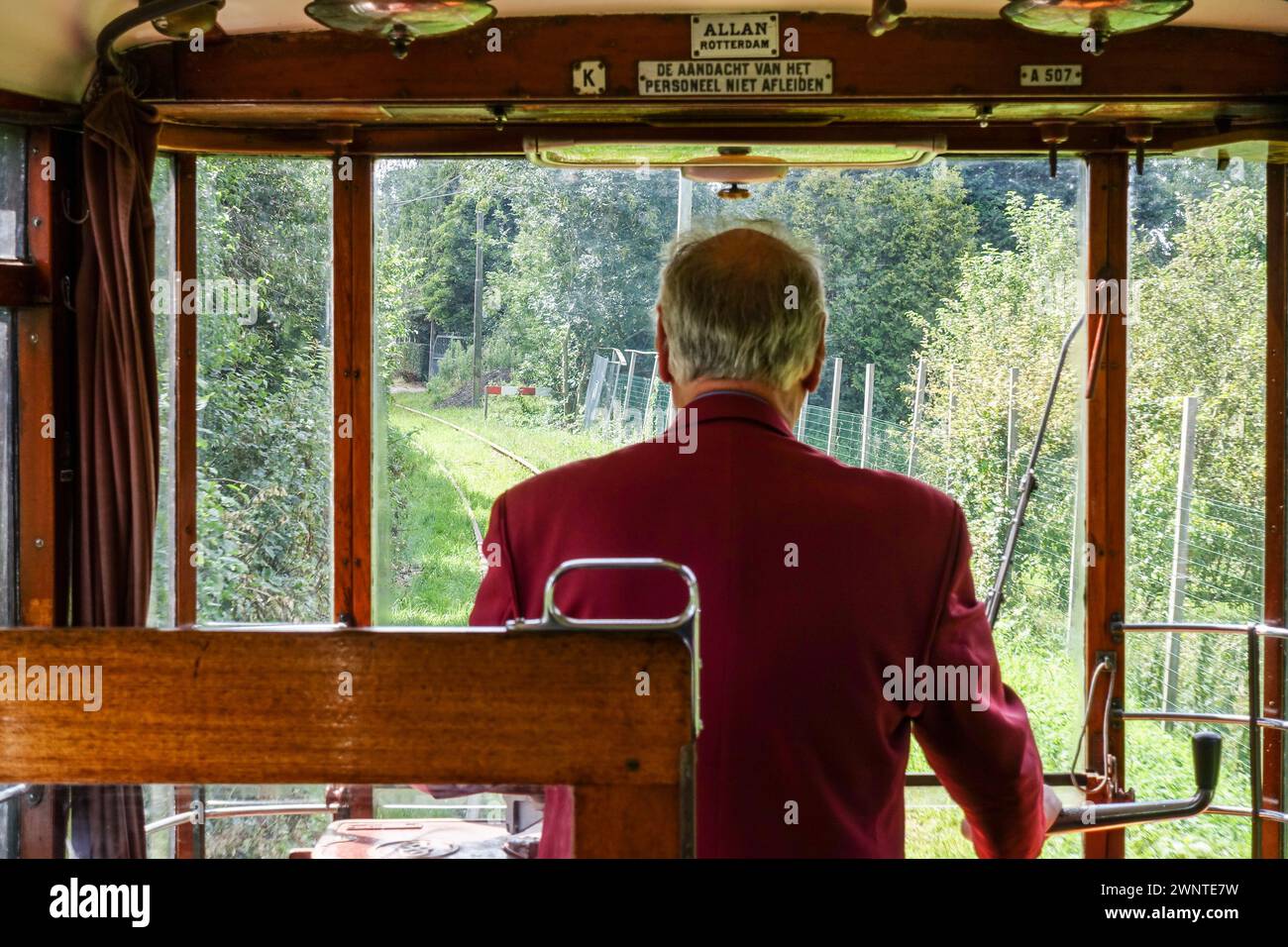 Straßenbahnführer in roter Jacke mit Blick aus der Kabine einer alten Straßenbahn, die malerische Eisenbahnstrecke und Bäume in Amsterdam, Niederlande, beobachtet Stockfoto