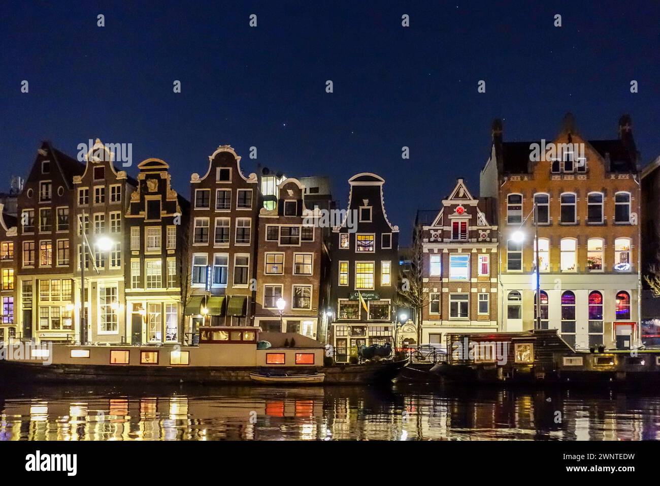 Historische holländische Gebäude, die bei Nacht entlang eines ruhigen Kanals in Amsterdam beleuchtet werden und sich auf der Wasseroberfläche spiegeln. Stockfoto