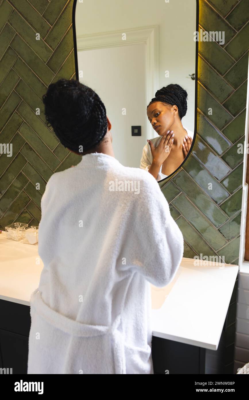 Eine junge afroamerikanische Frau in einem weißen Gewand untersucht ihr Spiegelbild im Badezimmer Stockfoto