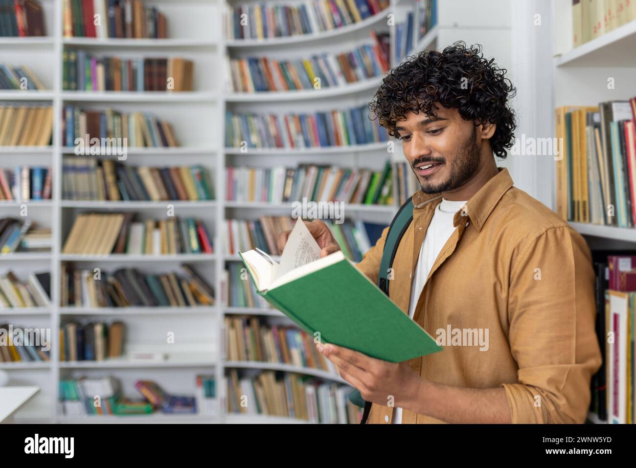 Ein erwachsener Mann steht in einer gut beleuchteten Bibliothek und liest ein grünes Buch. Bücherregale mit verschiedenen Büchern umgeben ihn, was eine ruhige Atmosphäre zum Studium und zur Literaturforschung nahe legt. Stockfoto Ein erwachsener Mann steht in einer gut beleuchteten Bibliothek und liest ein grünes Buch. Bücherregale mit verschiedenen Büchern umgeben ihn, was eine ruhige Atmosphäre zum Studium und zur Literaturforschung nahe legt. Stockfoto