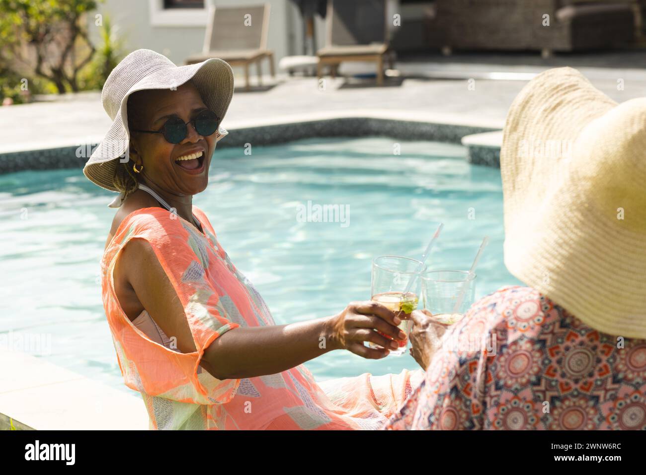 Eine ranghohe Afroamerikanerin in einem Sonnenhut lacht mit einer rassistischen Frau am Pool, mit Kopierraum Stockfoto
