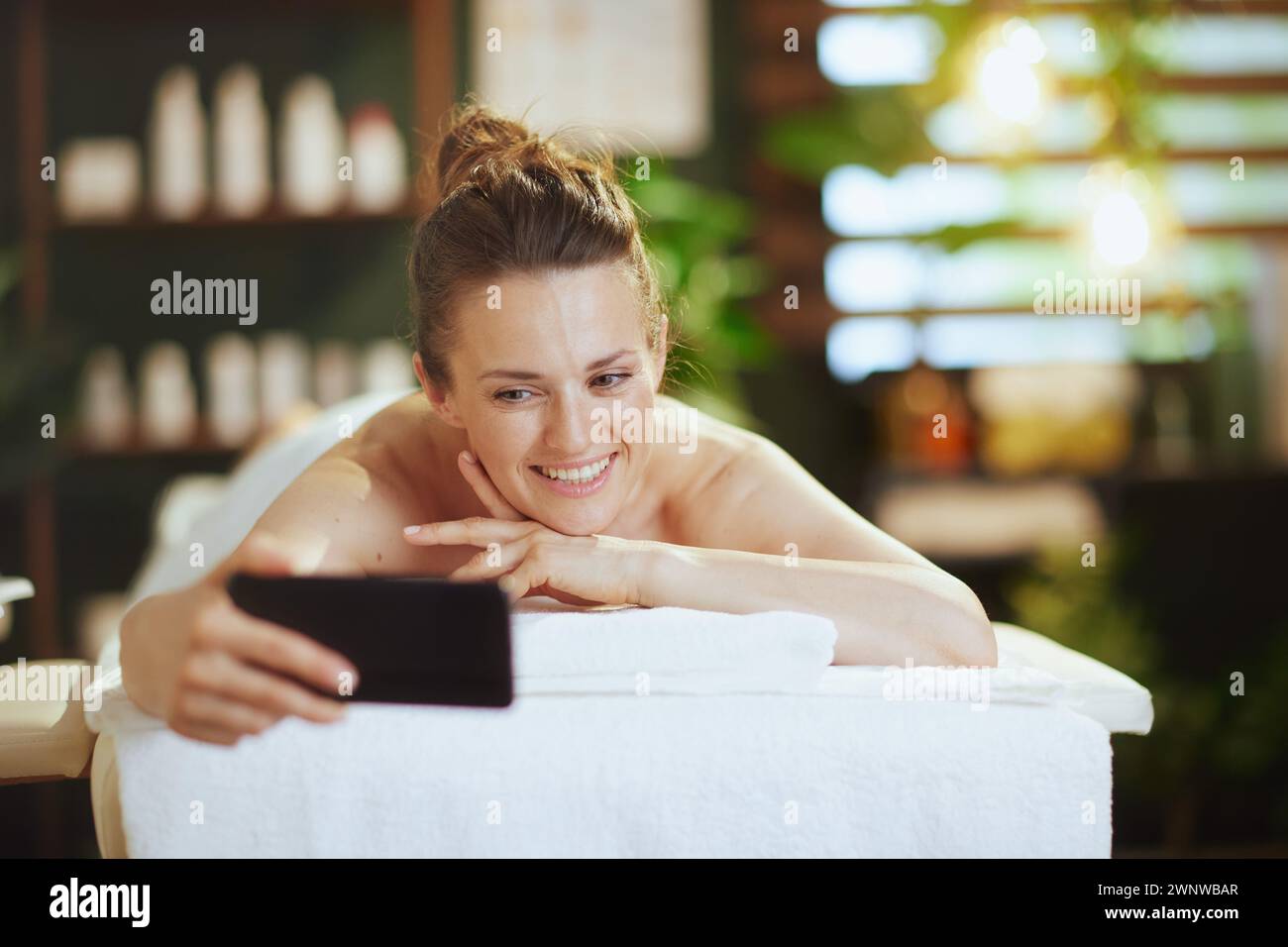 Gesundheitszeit. Lächelnde, moderne Frau im Spa-Salon, die Selfie macht und auf dem Massagetisch liegt. Stockfoto