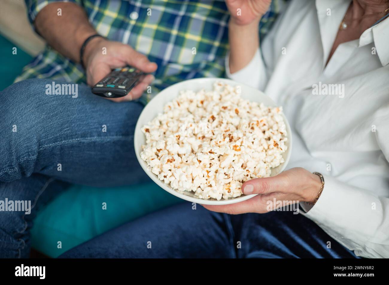 In einer Nahaufnahme halten die ineinander verwobenen Hände eines Paares liebevoll eine Fernbedienung und eine Schüssel Popcorn, die für einen gemütlichen Filmabend zusammen bereit sind. Dies in Stockfoto