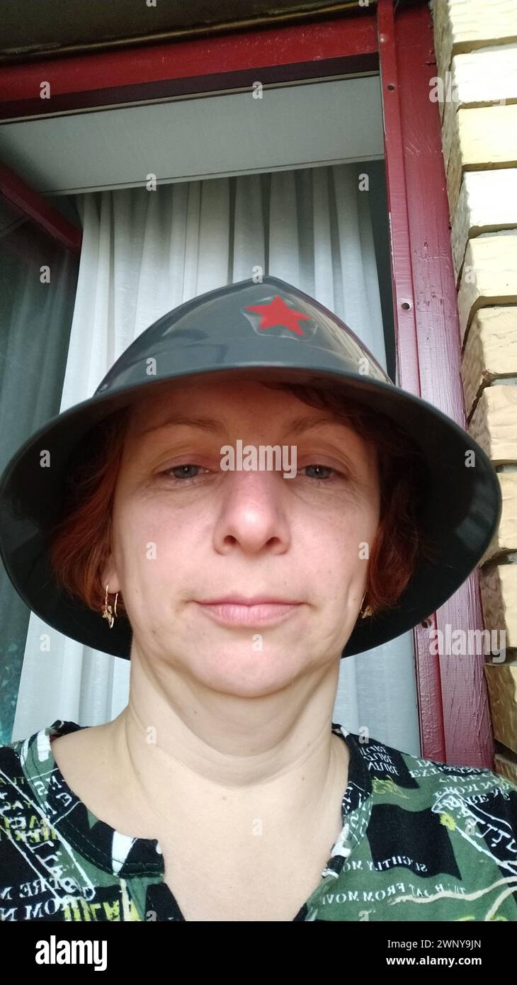 Eine 40-45-jährige Frau in einem grauen Feuerhelm mit einem roten Stern auf der Stirn macht ein Selfie. Halb lächeln, doppeltes Kinn und schlaffe Haut um den Hals Stockfoto