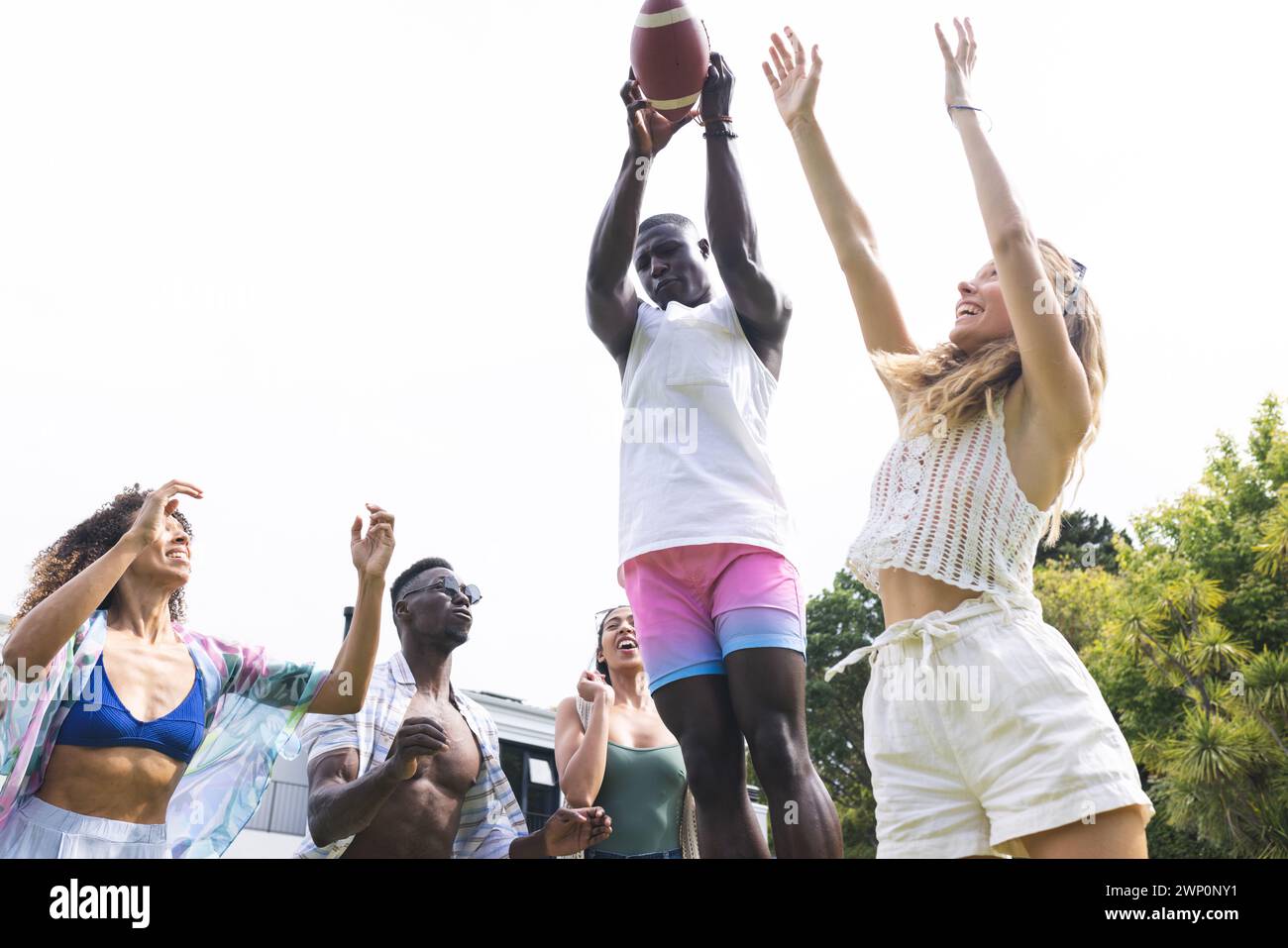 Ein junger Afroamerikaner fängt einen Fußball, umgeben von Freunden in legeren Sommerbekleidung Stockfoto