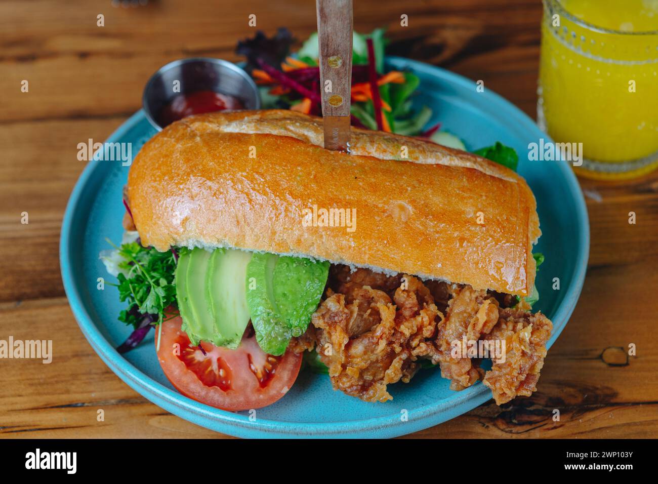 Ein Sandwich mit Avocado und Tomate auf einem blauen Teller. Ein Messer steht aus dem Sandwich Stockfoto