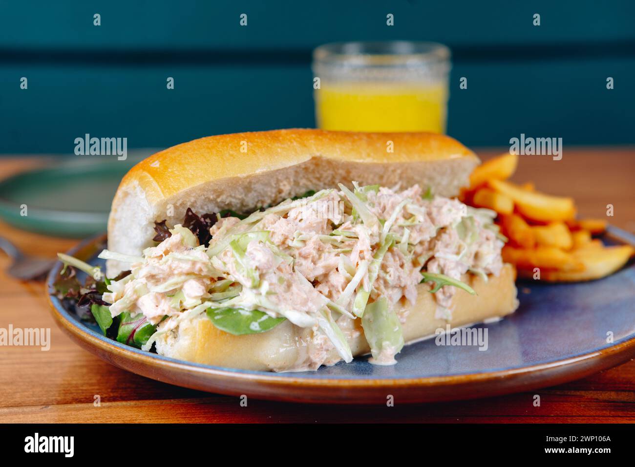 Ein Sandwich mit Salat und Huhn liegt auf einem Teller neben einem Glas Orangensaft. Das Sandwich wird in zwei Hälften geschnitten und auf eine blaue Platte gelegt Stockfoto