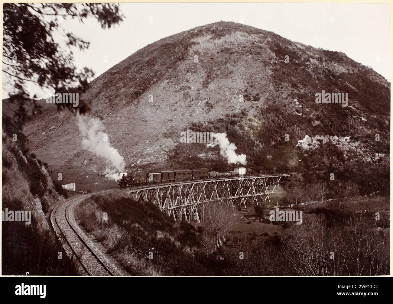 Dampfzug fährt über eine Holzbrücke vor einem großen Hügel. Lokomotiven der G-Klasse an beiden Enden des Zuges. Eisenbahnbrücke, südlich von Picton. Neuseeland um 1900 Stockfoto