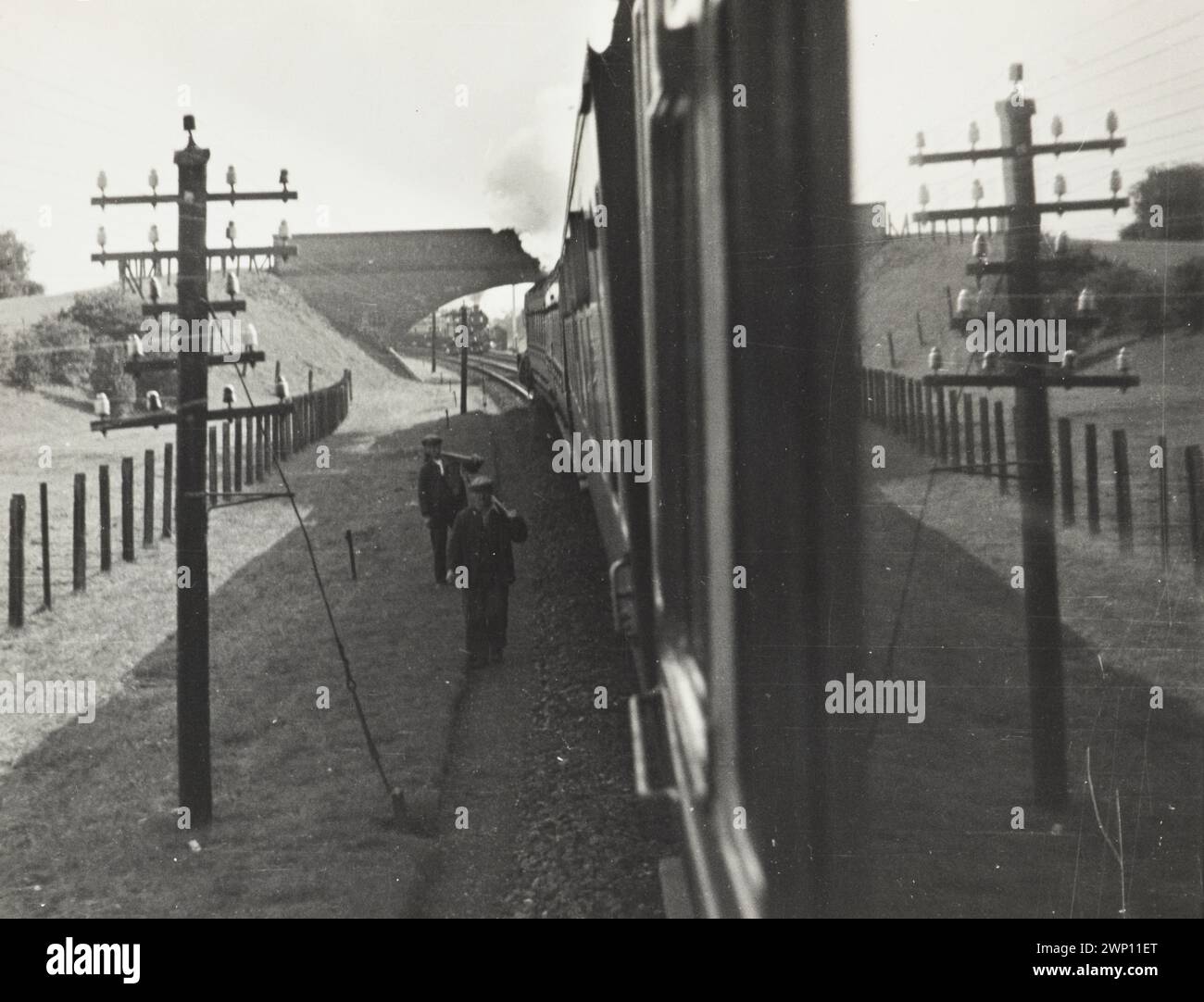 Überlegungen über einen Zug, England. 1930. Blick auf Zugwagen und auf eine Überbrücke. Ein Strommasten befindet sich im linken Vordergrund, während ein Zug, der auf dem anderen Gleis fährt, durch die Überbrücke zu sehen ist. Zwei Männer laufen mit ihren Werkzeugen an die Seite der Gleise. Stockfoto